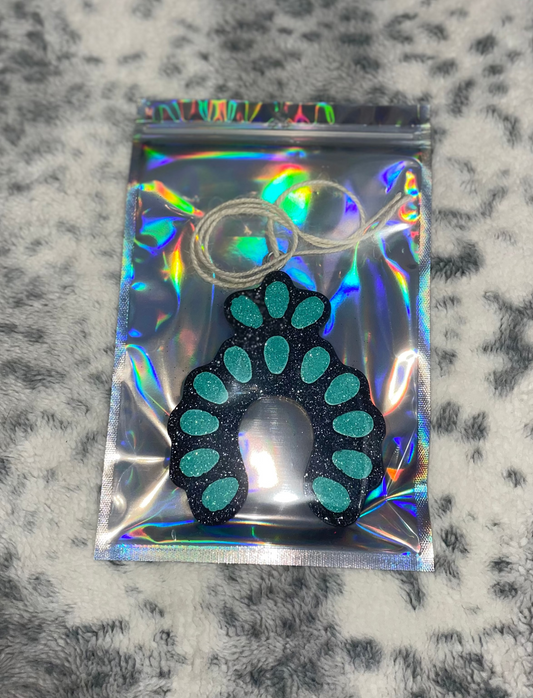 A turquoise and black air freshener with glitter, packaged in a plastic bag, with a textured background.
